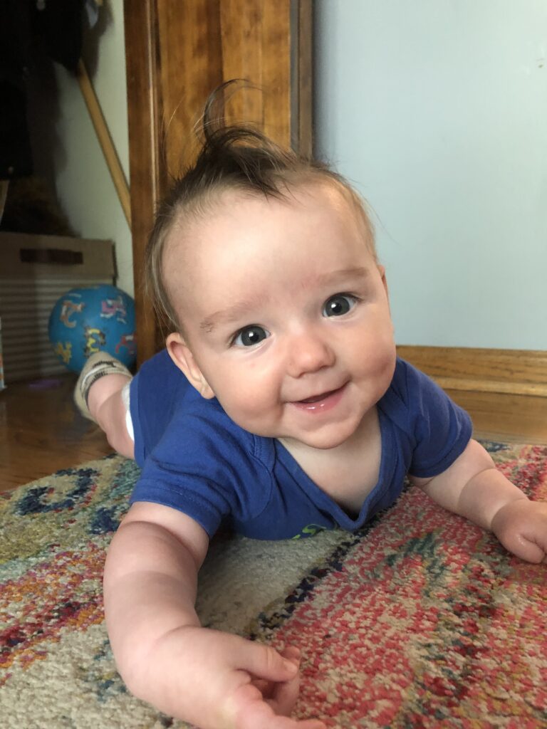 baby in blue onesie laying on belly, on colorful rug smiling, with playground ball in background