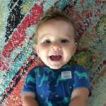 4 month old baby smiling wearing a dinosaur shirt laying on a colorful rug