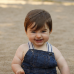brown haired 11 month old in overalls sitting in sand with a funny expression