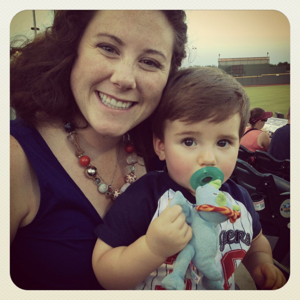 A women with a chunky necklace, holding a one year old at a baseball game. The one year old has a dinosaur Wubbanub pacifier in his mouth.