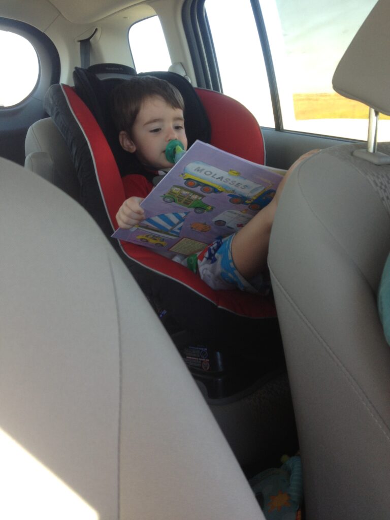 A one year old in the car, in a car seat, reading a book with a pacifier in his mouth