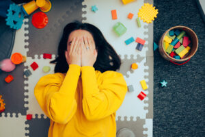 woman with dark hair in yellow sweater laying on the ground covering her face, surrounded by toys