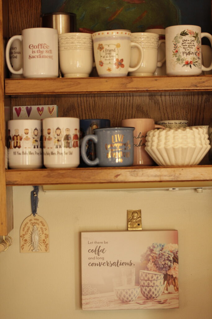 coffee mugs on shelves with a sign that says "Let there be coffee and long conversations" A relic of St. Padre Pio and a sign that says, "someone we love plays in Mary's garden above."