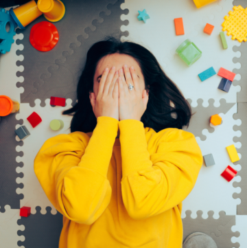 woman in bright yellow sweater laying on the ground surrounded by toys with her hands over her face